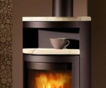 The image shows a section of a wood-burning stove, specifically the upper part of the "Troll III" model made of black steel. The focus is on a detail of the stove with an elegantly curved storage compartment on which a cup is standing. The viewing window below shows a lively play of flames, creating a cozy and inviting atmosphere. The sturdy steel body emphasizes the high-quality workmanship of the wood-burning stove. Ideal for stylish heating solutions in a modern design.