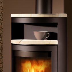 The image shows a section of a wood-burning stove, specifically the upper part of the "Troll III" model made of black steel. The focus is on a detail of the stove with an elegantly curved storage compartment on which a cup is standing. The viewing window below shows a lively play of flames, creating a cozy and inviting atmosphere. The sturdy steel body emphasizes the high-quality workmanship of the wood-burning stove. Ideal for stylish heating solutions in a modern design.