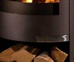 The image shows a section of a wood-burning stove with a focus on the wood compartment. At the bottom of the image, you can see logs neatly stacked. The wood compartment complements the wood-burning stove, which has an elegant steel design and a modern black finish. This combination creates a cozy, warm atmosphere in any room.