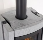 The image shows a section of an elegant wood-burning stove. The focus is on the top cover, which is made of gray stone. This robust stone surface gives the stove a modern and stylish look. The black smoke vent contrasts sharply with the light-colored stone. This wood-burning stove is ideal for modern living spaces where design and functionality are key.