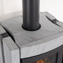 The image shows a section of an elegant wood-burning stove. The focus is on the top cover, which is made of gray stone. This robust stone surface gives the stove a modern and stylish look. The black smoke vent contrasts sharply with the light-colored stone. This wood-burning stove is ideal for modern living spaces where design and functionality are key.