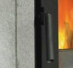 De afbeelding toont een detail van een houtkachel met een duidelijke focus op de deur en de handgreep. De vurige vlammen op de achtergrond stralen een uitnodigende warmte uit en de massieve handgreep zorgt voor een veilige bediening. De houtkachel is gedeeltelijk bekleed met steen, wat het ontwerp een elegante en stijlvolle uitstraling geeft. Perfect voor een moderne woonomgeving en gezellige avonden voor de houtkachel.
