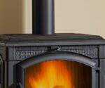 The picture shows a section of a stove with the focus on the top and the front. The close-up highlights the artistically designed metalwork. A warm, lively fire is visible through the viewing window. This wood-burning stove combines classic design with modern, efficient combustion technology. Perfect for creating a cozy atmosphere at home.