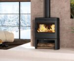 Image description: An elegant black wood-burning stove stands in a modern room, in front of a large window with a view of a snow-covered landscape. A lively fire blazes inside the stove, creating a cozy warmth and atmosphere. Underneath the stove is a wood compartment, which is stocked with firewood. This wood-burning stove contributes to a stylish and cozy living atmosphere.