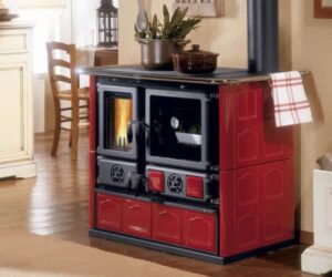 An elegant stove with a pink, border-reinforced design. The stove has two doors: one with a window that provides a view of the crackling fire and another door that may serve as a warming compartment. It stands on a wooden floor in a cozy room with some furniture in the background. A perfect wood-burning stove that brings both warmth and charm to any home. Ideal for lovers of traditional wood-burning stove design.
