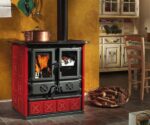 A rustic wood-burning stove in a classic design stands in a cozy room. The stove has striking red tile cladding and two visible doors, one of which offers a view of the blazing fire. The wood-burning stove radiates warmth and coziness, while a copper pot stands on top of the stove. In the background, wooden furniture and a basket of firewood can be seen, emphasizing the inviting atmosphere. Ideal for lovers of classic wood-burning stoves and those who want to enhance their surroundings with a stylish wood-burning stove.