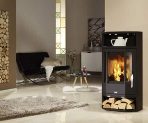 Ein eleganter Kaminofen im modernen Design steht in einem gemütlichen Raum. Der Ofen aus Stahl hat eine große Glasscheibe, durch die helle Flammen sichtbar sind, was eine warme und einladende Atmosphäre schafft. Darunter befindet sich ein praktisches Holzfach, in dem Holzscheite ordentlich gelagert sind. Der Kaminofen ist ein stilvoller Mittelpunkt des Raumes, der sowohl funktional als auch ästhetisch ansprechend ist. Ideal für ein behagliches Wohnambiente mit einem effizienten Holzofen.