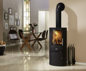 This picture shows a stylish steel wood-burning stove in a modern living environment. The stove, which takes center stage, has a clear, large glass panel through which a living fire can be seen. Next to the stove are discreet fireplace accessories that emphasize the functional elegance of the room. The warm glow of the wood-burning stove creates a cozy atmosphere in the room. A table with chairs and decorative elements can be seen in the background, harmoniously complementing the room.