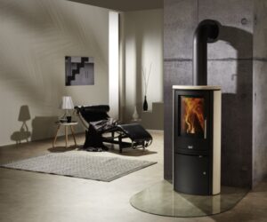 The picture shows an elegant cream-colored wood-burning stove of the "Viva" model in a modern living room. The wood-burning stove stands in front of a gray wall and has a generous glass front through which a lively play of flames is visible. The stove is placed on a transparent base plate, which is stylishly integrated into the minimalist interior. Modern furniture and decorative elements can be seen in the background. The wood-burning stove not only brings warmth to the room, but is also an aesthetic highlight. Keywords: wood-burning stove, wood-burning stove, modern, cream.