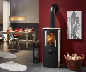A stylish stove with a sandstone cladding stands in a modern living room. The stove is matt black with a large, arched viewing window showing a living fire. Next to the stove is a basket of logs and in the background is a dining area with a long table. The contrast between the warm sandstone and the red background gives the room a cozy atmosphere. Perfect for the cozy warmth of a wood-burning stove.