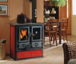 A stylish wood-burning stove with red cladding and a classic design stands in a cozy living room. A pot is placed on the stove's cooking surface. The fire in the combustion chamber is clearly visible and radiates a warm atmosphere. In the background, you can see a dining area with a wooden table and chairs. This wood-burning stove combines functionality with appealing aesthetics, ideal for a cozy home.