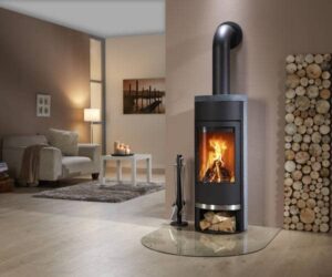 Ein stilvoller Kaminofen mit elegantem Speckstein in einem modernen Wohnzimmer. Der Ofen, der auf einer schützenden Glasplatte steht, hat ein einladendes, leuchtendes Feuer im Inneren, das Wärme und Gemütlichkeit ausstrahlt. Neben dem Ofen ist ein Holzlager mit ordentlich gestapeltem Brennholz zu sehen. Ideal für jene, die ein funktionales und ansprechendes Heizgerät suchen.