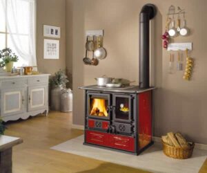 Ein stilvoller Kaminofen in einem gemütlichen Raum. Der Ofen ist in einem eleganten Rot gehalten und verleiht der Umgebung eine warme Atmosphäre. Links oberhalb sieht man die Brennkammer mit einer klaren Scheibe, durch die das lodernde Feuer sichtbar ist. Neben dem Ofen befindet sich ein offenes Holzfach, in dem Feuerholz ordentlich gestapelt ist. Der Kaminofen kombiniert traditionelle Funktionalität mit einem modernen Design und ist sowohl ein praktisches Heizgerät als auch ein optisches Highlight für den Wohnraum.