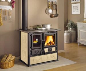 A stylish wood-burning stove stands in a cozy room. The stove has cream-colored cladding and a visible fire door through which warm flames can be seen. To the left is a second door, possibly for wood or ash removal. Cookware is placed on top of the stove, emphasizing its multifunctional use as a wood-burning stove and cooking area. Kitchen utensils hang on the wall, accentuating the ambiance of a welcoming, rustic home. This wood-burning stove combines aesthetics with functionality and creates a cozy atmosphere.