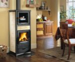 The picture shows an elegant wood-burning stove in a classic design with light-colored decorations and a black body. This wood-burning stove, called "Rossella Plus Forno", has an integrated baking compartment, which can be seen at the top of the stove. The large glass door provides an atmospheric view of the flickering fire, while a separate compartment underneath offers space for wood. The stove is set up in a cozy living space that is inviting and warm. With its practical and aesthetic design, this wood-burning stove is an attractive addition to any living area. Keywords: wood-burning stove, wood-burning stove, wood-burning stove with baking compartment.