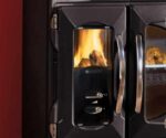 The image shows a detailed section of a wood-burning stove, specifically the glass viewing panel. The panel allows you to see the lively flames of the wood-burning stove, which creates a cozy atmosphere. The shiny metal handles and stylish black design emphasize the modern aesthetics of the stove. Perfect for enjoying the warmth and atmosphere of a wood-burning stove.