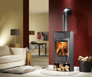 An elegant stove made of black steel stands in a modern living room. The fire blazes behind a large glass panel, giving the room a warm and inviting atmosphere. Below the combustion chamber is a spacious wood compartment that displays neatly stacked wood. The stove is placed against a red wall, which creates a stylish contrast. This wood-burning stove is a perfect addition to any home looking for a cozy heat source.