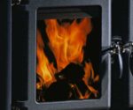 De afbeelding toont een gedetailleerd fragment van een houtkachel, met de nadruk op de vuurplaat. Achter de glasplaat laaien levendige vlammen op, die een warme en uitnodigende sfeer creëren. De robuuste metalen rand van de houtkachel geeft het ontwerp een stijlvolle en tijdloze uitstraling. Ideaal als aanvulling op een gezellige woonkamer met een houtkachel.