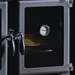 The image shows a detail of a wood-burning stove, focusing on a thermometer and a small stove window. The stove has a robust and functional appearance, perfect for anyone who wants a reliable wood-burning stove in their home. This detail highlights the high-quality workmanship and elegant design, while the thermometer allows for precise temperature control—ideal for efficient heating with a wood-burning stove.