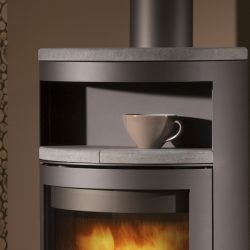 De afbeelding toont het bovenste deel van een moderne houtkachel, met name het gedeelte boven de glazen deur, waar een grijze mok op een plank staat. De kachel is gemaakt van staal en gietijzer, wat hem een elegant en eigentijds design geeft. Door de glazen deur schijnt een rustgevend vlammenspel. Deze kachel is perfect als stijlvolle aanvulling voor elke woonkamer en creëert een gezellige sfeer. Ideaal voor iedereen die op zoek is naar een hoogwaardige houtkachel voor zijn huis.