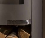 De afbeelding toont een detail van een grijze stalen kachel met speciale aandacht voor het onderste gedeelte. De solide afwerking en het moderne ontwerp zijn duidelijk zichtbaar. Het detail benadrukt de afgeronde randen en de elegante overgang tussen de materialen. Op de achtergrond zijn houtblokken te zien, die een gevoel van warmte en gezelligheid uitstralen. Dit detail van de houtkachel illustreert de harmonieuze combinatie van design en functionaliteit in een moderne kachel.