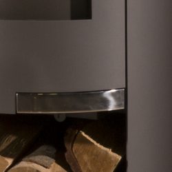 The image shows a section of a gray steel stove with a particular focus on the lower area. The solid workmanship and modern design are clearly visible. The section highlights the rounded edges and the elegant transition between the materials. Logs can be seen in the background, conveying a feeling of warmth and comfort. This section of the wood-burning stove illustrates the harmonious combination of design and functionality in a modern stove.
