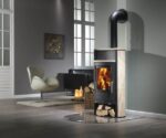 Een stijlvol ingerichte woonkamer met een centrale houtkachel. Het model "Piro 7" is voorzien van een elegante zandstenen bekleding, die de ruimte een natuurlijke en warme uitstraling geeft. Het vuur achter het glas zorgt voor een gezellige sfeer. De kachel heeft een handig houtvak aan de onderkant, waarin houtblokken netjes zijn opgestapeld. Een moderne, gebogen pijp voert de rook naar boven af.