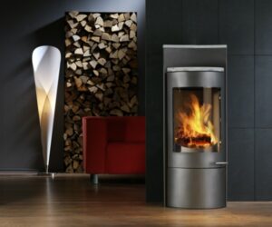 A stylish gray wood-burning stove can be seen in a modern living room. The stove has a large viewing window through which lively flames are visible, creating a warm and cozy atmosphere. In the background is a shelf with decoratively stacked firewood and a red sofa, giving the room an elegant and inviting ambience. The wood-burning stove blends harmoniously into the modern design of the surroundings and provides cozy warmth. An ideal example of efficient and aesthetic room heating with a wood-burning stove.