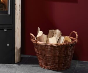 Dieses Bild zeigt einen robusten Weidenkorb, der mit Holzscheiten befüllt ist. Der Korb befindet sich auf einem dunklen Boden vor einer rot gestrichenen Wand und neben einem modernen Kaminofen. Der Holzkorb ist ein praktisches Zubehör für jeden Holzofen oder Kaminofen und ergänzt die gemütliche Atmosphäre in Wohnräumen wunderbar.