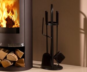 Das Bild zeigt ein elegantes Kaminbesteck-Set neben einem modernen Kaminofen. Der Ofen aus Metall hat eine große, gewölbte Scheibe, durch die ein lebendiges Feuer sichtbar ist, und ein praktisches Holzfach darunter, das mit Holzscheiten gefüllt ist. Das schwarze Kaminbesteck besteht aus einem Ständer mit Besen, Schaufel, Haken und Schürhaken, ideal für die Pflege eines Holzofens. Diese Gestaltung verleiht jedem Raum eine stilvolle und funktionale Atmosphäre.