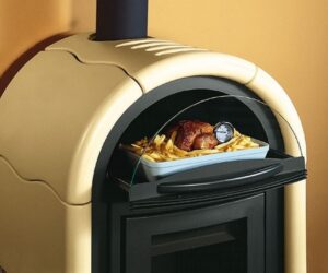 Alt text: The image shows a section of a wood-burning stove with a focus on the baking compartment. The cream-colored wood-burning stove has a stylish, curved design. The baking compartment contains a roast in a baking dish, accompanied by French fries and a thermometer for temperature control. The wood-burning stove combines design and functionality in an attractive element for any home.