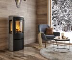 Een stijlvolle houtkachel staat in een modern ingerichte woonkamer met houten wanden en een groot raam dat uitzicht biedt op een besneeuwde buitenruimte. De kachel heeft een strak design en een glazen voorkant waardoor de vlammen zichtbaar zijn. Er staat een grijze fauteuil met een kussen en een deken naast, terwijl een rond bijzettafeltje met een kopje erop op een zacht tapijt staat. Deze houtkachel geeft de kamer een gezellige sfeer en nodigt uit tot ontspanning. Perfect voor iedereen die op zoek is naar een elegante en efficiënte open haard.
