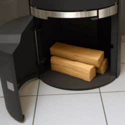 De afbeelding toont het geopende houtmagazijn van een moderne houtkachel, model Filou II HL. In de houtkachel bevinden zich meerdere houtblokken. De constructie is van hoge kwaliteit, met strakke lijnen en een elegante zwarte afwerking. Bijzonder opvallend is het praktische houtvak, dat zorgt voor een eenvoudige opslag van brandhout direct bij de houtkachel. Ideaal voor een efficiënte verwarmingsoplossing thuis.