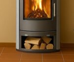 The picture shows a wood-burning stove. The stove has a large, curved glass door through which the fire can be seen. Below the fire chamber is a compartment for storing logs. This wood compartment is clearly visible and filled with several logs. The stove itself is dark in color and stands on a tiled floor. The walls in the background are a warm color. The picture focuses particularly on the wood compartment of the stove.