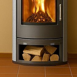 The picture shows a wood-burning stove. The stove has a large, curved glass door through which the fire can be seen. Below the fire chamber is a compartment for storing logs. This wood compartment is clearly visible and filled with several logs. The stove itself is dark in color and stands on a tiled floor. The walls in the background are a warm color. The picture focuses particularly on the wood compartment of the stove.