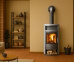 The picture shows an elegant, modern wood-burning stove in a cozy living room. The stove has a large glass door through which a lively fire can be seen, creating a warm and inviting atmosphere. There is a practical wood compartment under the firebox, which is filled with logs. Next to the stove is a decorative shelf wall with vases and plants as well as a cozy seating area, which make a harmonious addition. The stove blends seamlessly into the stylish design of the room and offers both functionality and aesthetic value.