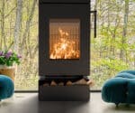Detailaufnahme eines schwarzen Kaminofens mit loderndem Feuer, daneben grüne Pflanzen und blaue Sessel.