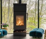 Detail eines schwarzen Kaminofens mit brennendem Feuer, im Hintergrund große Fenster mit Blick auf Bäume.