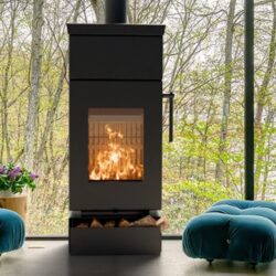 Detail eines schwarzen Kaminofens mit brennendem Feuer, im Hintergrund große Fenster mit Blick auf Bäume.