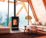 Kaminofen Thermorossi Zen W in modernem Wohnzimmer mit großen Fenstern, brennendem Feuer und gemütlicher Einrichtung. Holzlagerfach sichtbar.