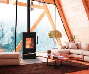 Kaminofen Thermorossi Zen W in modernem Wohnzimmer mit großen Fenstern, brennendem Feuer und gemütlicher Einrichtung. Holzlagerfach sichtbar.